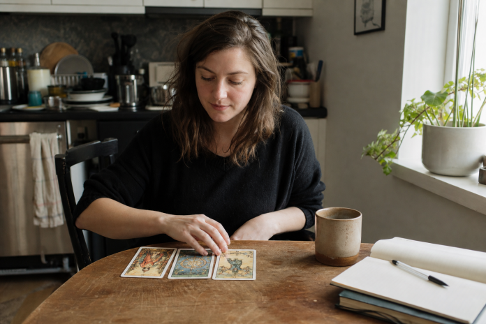 Woman in her thirties laying out three illustrated cards at a kitchen table beside a coffee mug and notebook in morning light