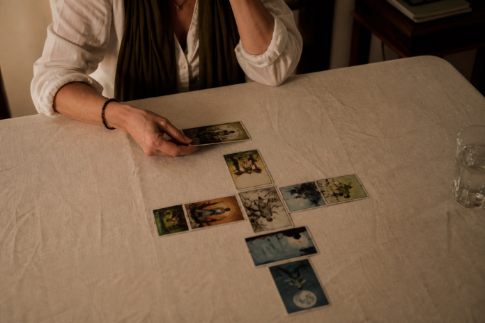 Tarot spreads: ten-card Celtic Cross layout on a dining table Overhead view of a woman placing the last card of a ten-card cross-shaped tarot spread on a linen-covered dining table in evening lamp light