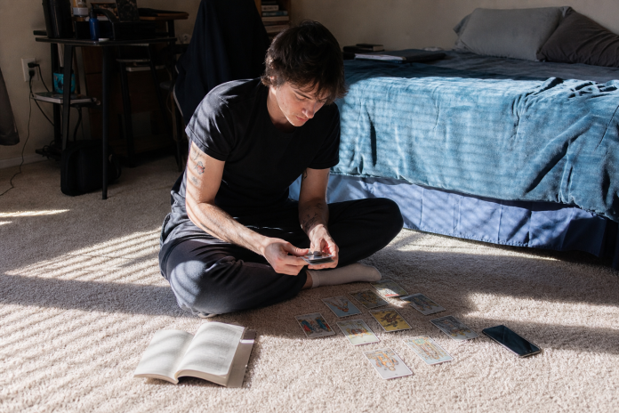 Tarot cards for beginners: learning a first deck on a bedroom floor Young man sitting cross-legged on a bedroom carpet with a beginner tarot deck spread out, an open guidebook and a phone beside him in afternoon light