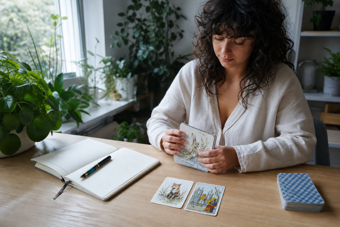 A woman at an oak desk compares an oracle card with a tarot card beside an open journal in a plant-filled home office