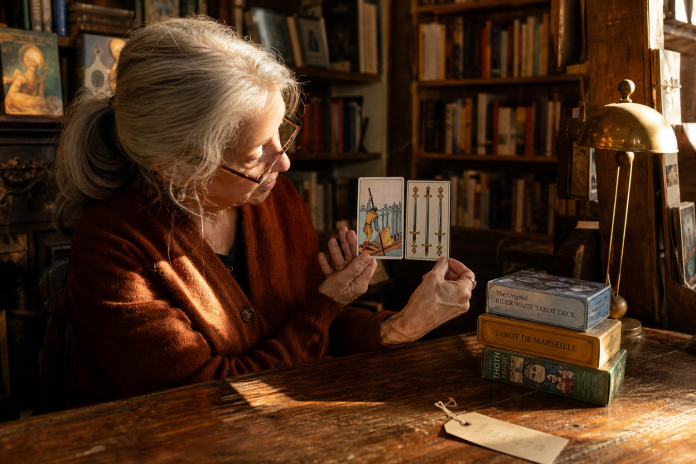 A woman in a bookshop compares an illustrated tarot card with a plain Marseille style card beside stacked deck boxes