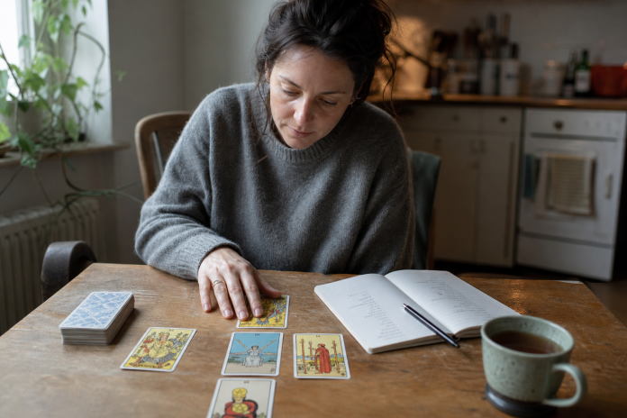 Woman at a kitchen table with a five-card tarot decision spread and a notebook with a pros-and-cons list