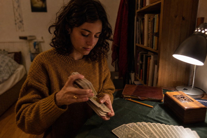 Young woman shuffling a worn tarot deck at a lamplit desk with a cloth, a wooden deck box and a journal