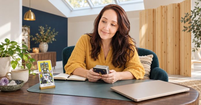 The Chariot tarot card in a reading Woman holding a tarot deck beside the Chariot card in a brass stand and a closed champagne-coloured laptop.