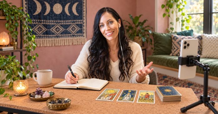 Preparing for a tarot session Dark-haired woman with an earbud and notebook beside tarot cards and a phone on a stand.