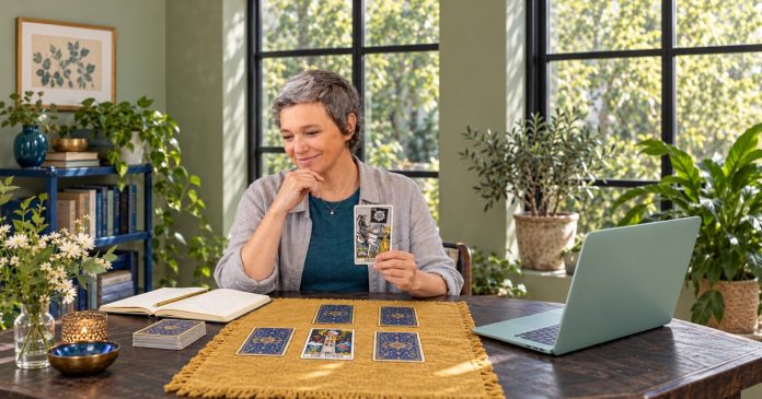 Six feared tarot cards — Death and the Tower A woman holds the Death tarot card beside a Tower card, four face-down cards, a notebook and a green laptop.