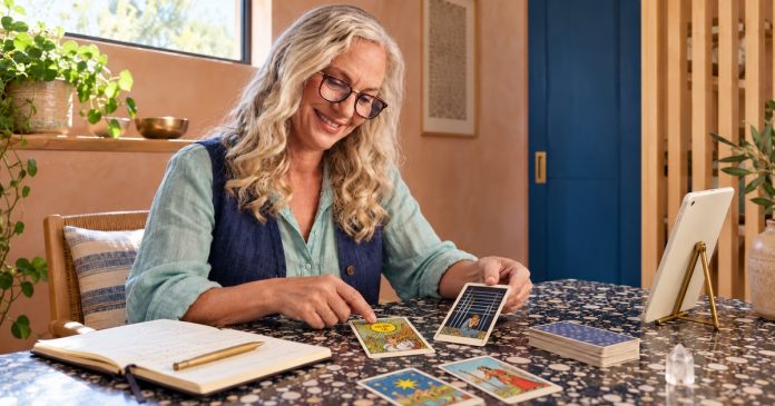 Reading color symbolism in tarot Silver-blonde woman with tortoiseshell glasses comparing bright and dark tarot cards at a terrazzo table beside a small ivory tablet.