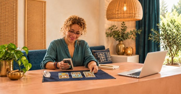 Woman with golden curls and round glasses arranging tarot cards beside a crystal, notebook and white laptop at a peach-colored table.